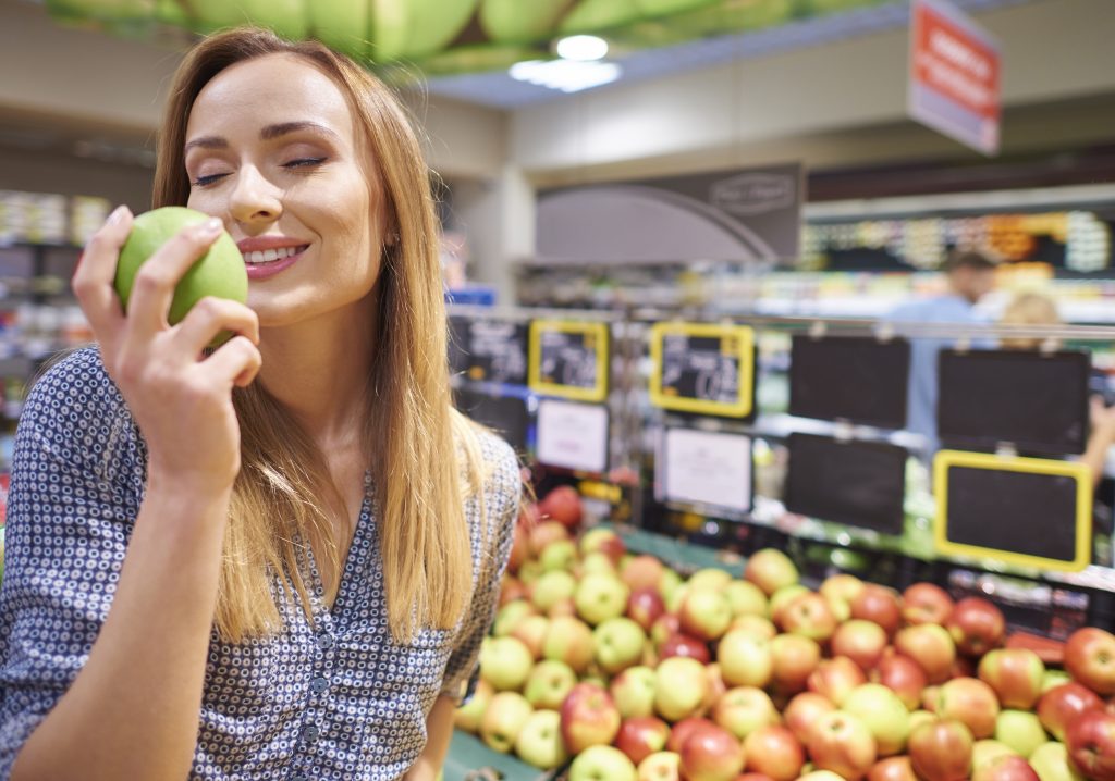 Uma mulher sorridente, com os olhos fechados, segurando uma maçã verde perto do rosto para cheirá-la. Ela está na seção de frutas de um supermercado, com uma grande variedade de maçãs e outras frutas ao seu redor.