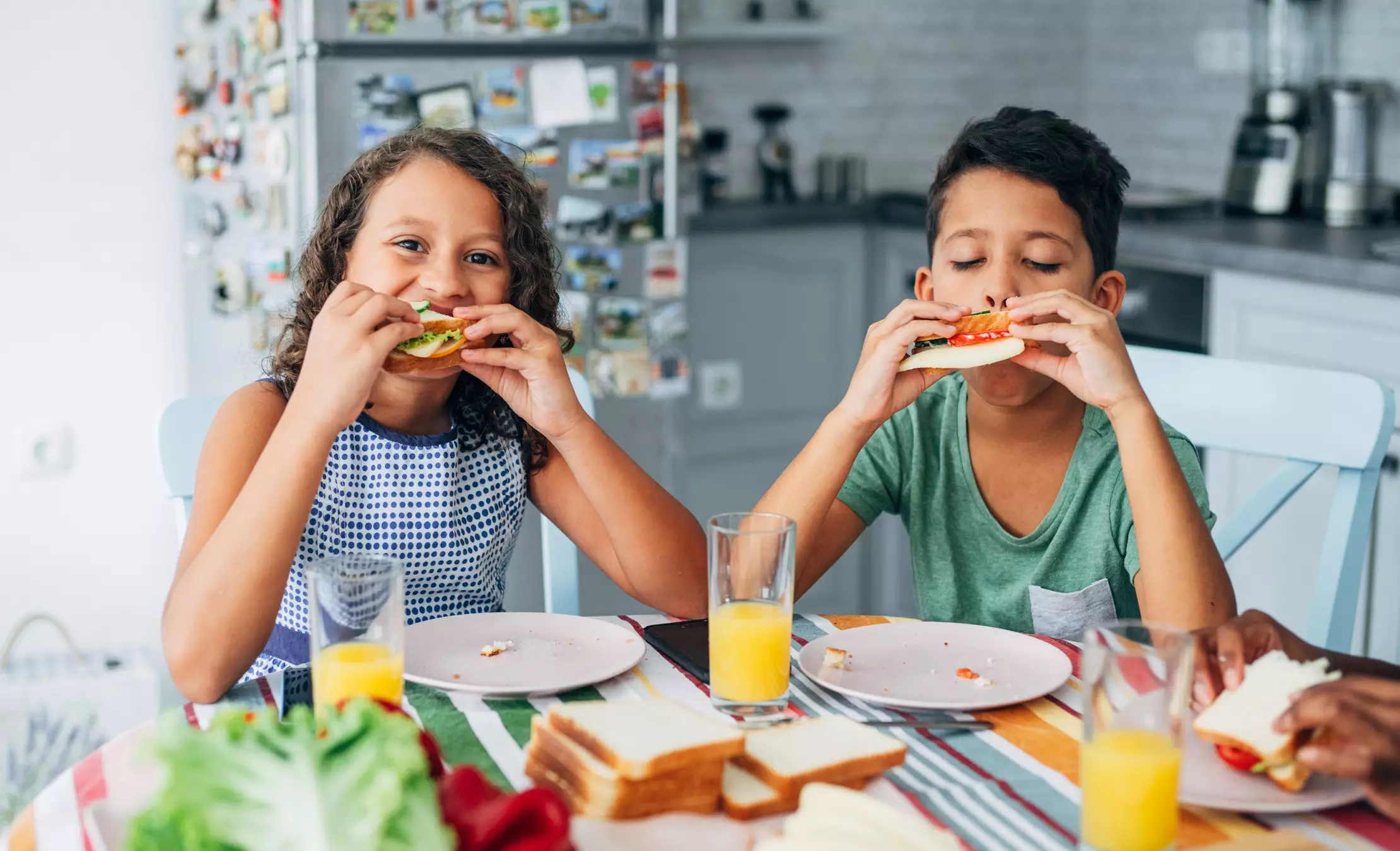 duas crianças estão mordendo um sanduíche enquanto estão sentadas à mesa; na mesa é possível ver pratos brancos, sucos de laranja, pães de forma e ao fundo uma cozinha.