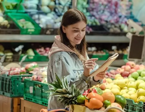 Uma mulher jovem, de cabelos castanhos e vestindo uma jaqueta jeans e um moletom rosa, está sorrindo enquanto usa o dedo indicador para checar itens em uma lista de compras. Ela está em um supermercado, em frente à seção de frutas e vegetais, onde abacaxis, laranjas, pimentões e maçãs estão à vista. O carrinho de compras dela está cheio de produtos frescos.