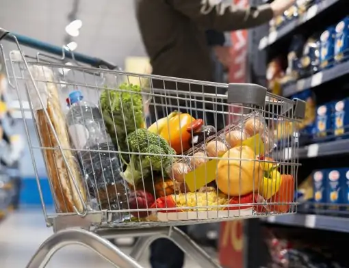 Carrinho de supermercado em destaque, lotado de compras como frutas, verduras, ovos, pão e uma garrafa de água, em um corredor de supermercado. O foco está no carrinho, com as prateleiras ao fundo levemente desfocadas e uma pessoa de costas.