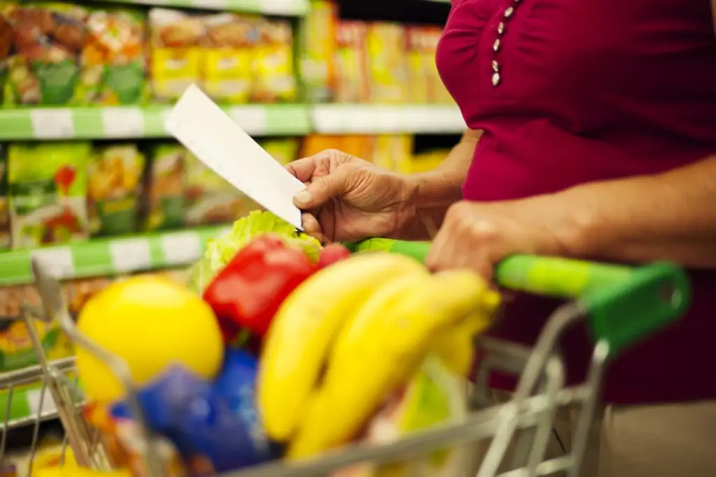 Mulher de meia-idade segurando um carrinho de compras enquanto olha uma lista de papel, com o carrinho cheio de frutas e vegetais frescos.